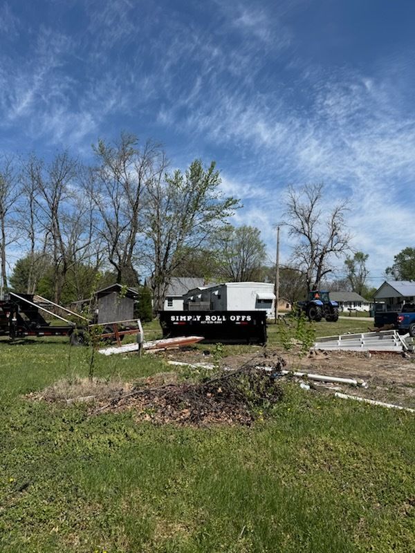 Storm-damaged yard with debris, a trailer, and fallen trees under a blue sky