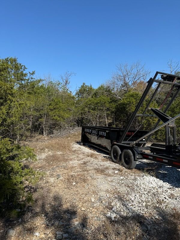 A black roll-off dumpster trailer parked on a gravel path near trees and brush on a sunny day.