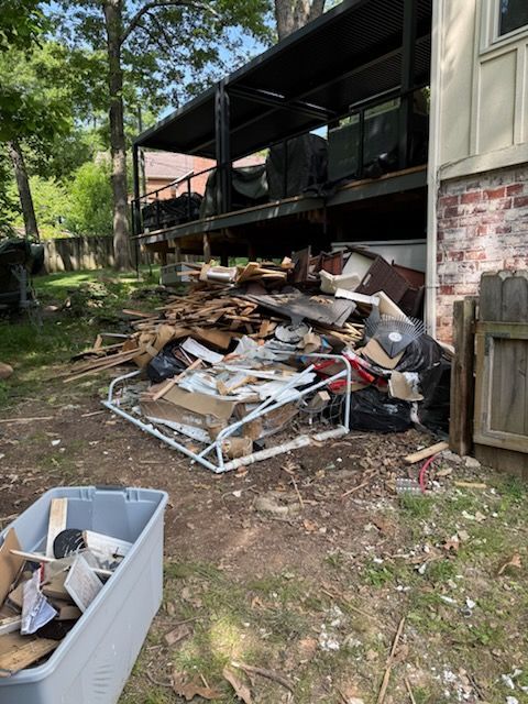 Outdoor debris pile with broken furniture and trash beside a house and deck, in a wooded yard.