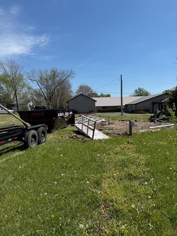 Rural yard with debris and a black trailer beside a house under a clear blue sky