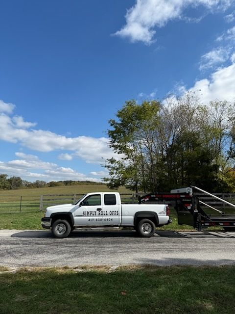 White pickup truck with a trailer parked on gravel, under a blue sky. Rural setting with a fence and trees.