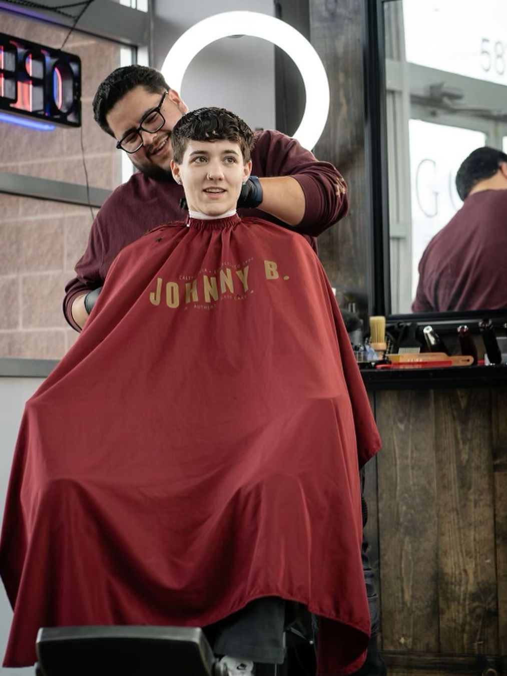 A man is getting his hair cut by a barber wearing a red cape that says johnny b.