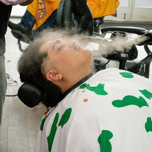 A man in a barber chair receiving a facial steam. Steam covers his face; he wears a cape.
