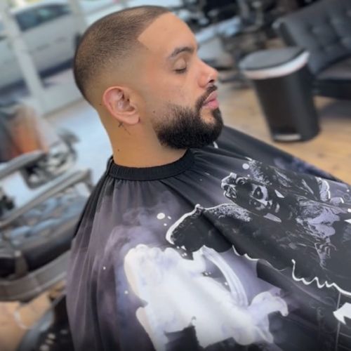 Man in barber cape getting haircut, faded hair, beard. Indoors.