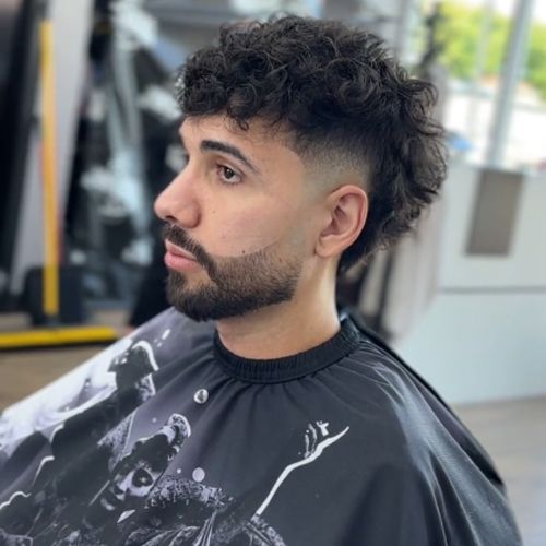 Man with curly hair and beard in barber cape, side profile. Fade haircut.