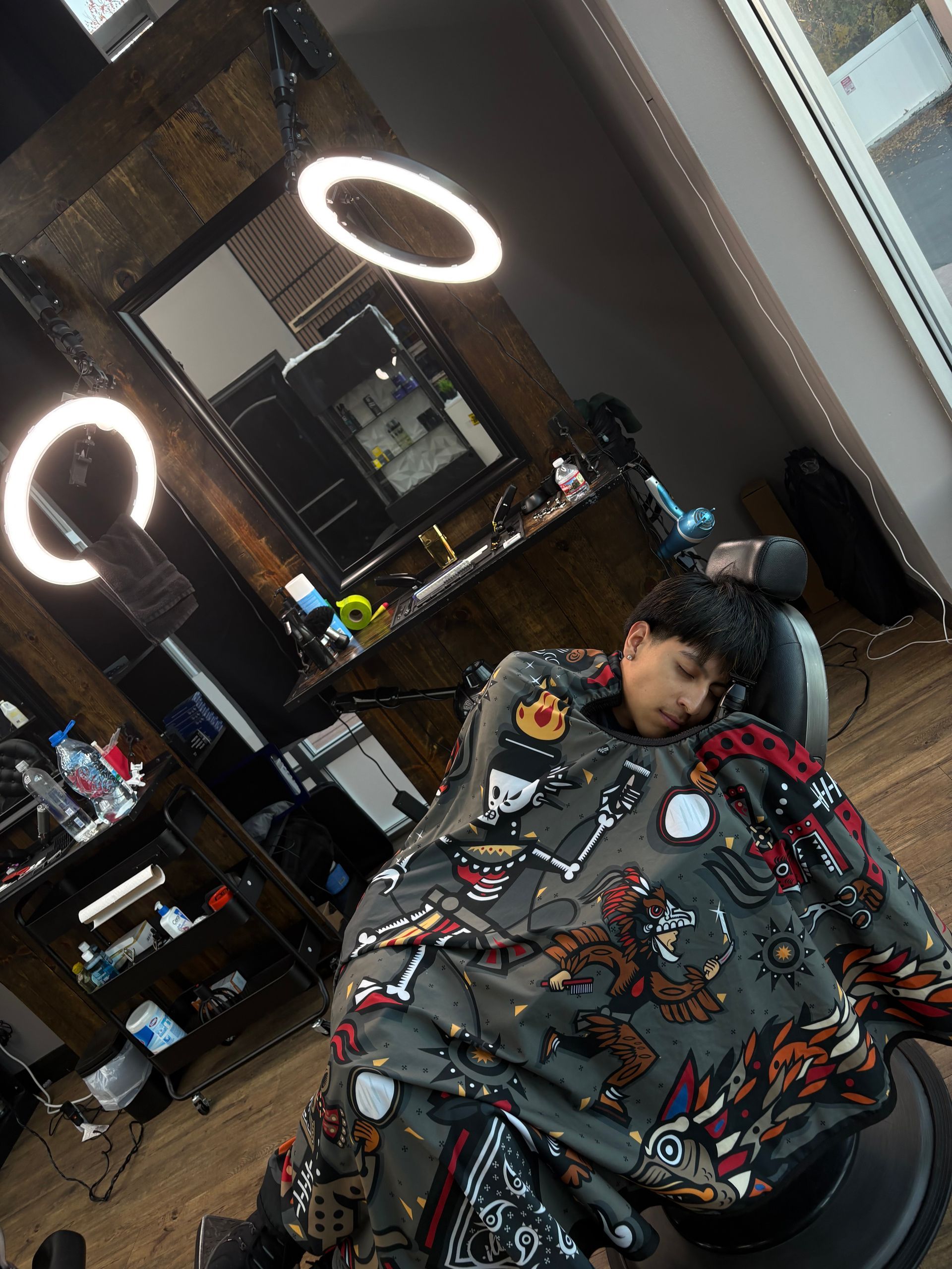 Person asleep in barber chair, wearing patterned cape. Barber shop interior with ring lights and mirror.