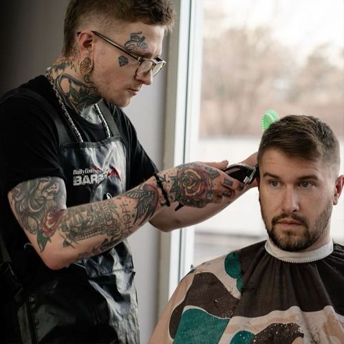 Barber with tattoos cutting a man's hair with clippers near a window.