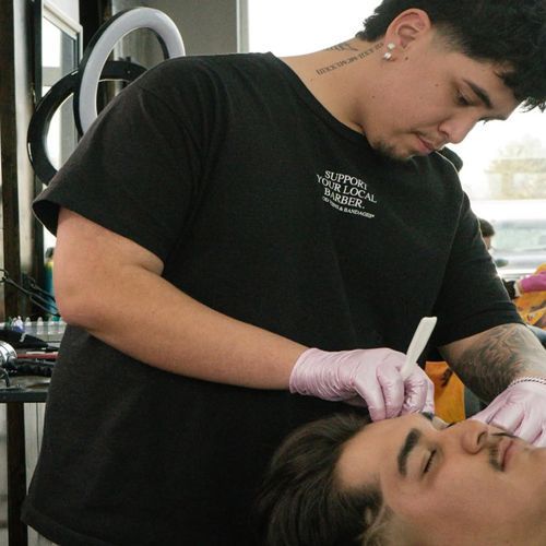 Barber shaping client's eyebrow. Barber is wearing a black shirt and pink gloves.