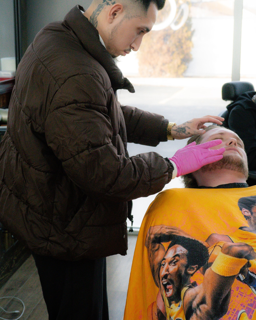 A man with a tattoo on his neck is shaving another man 's face
