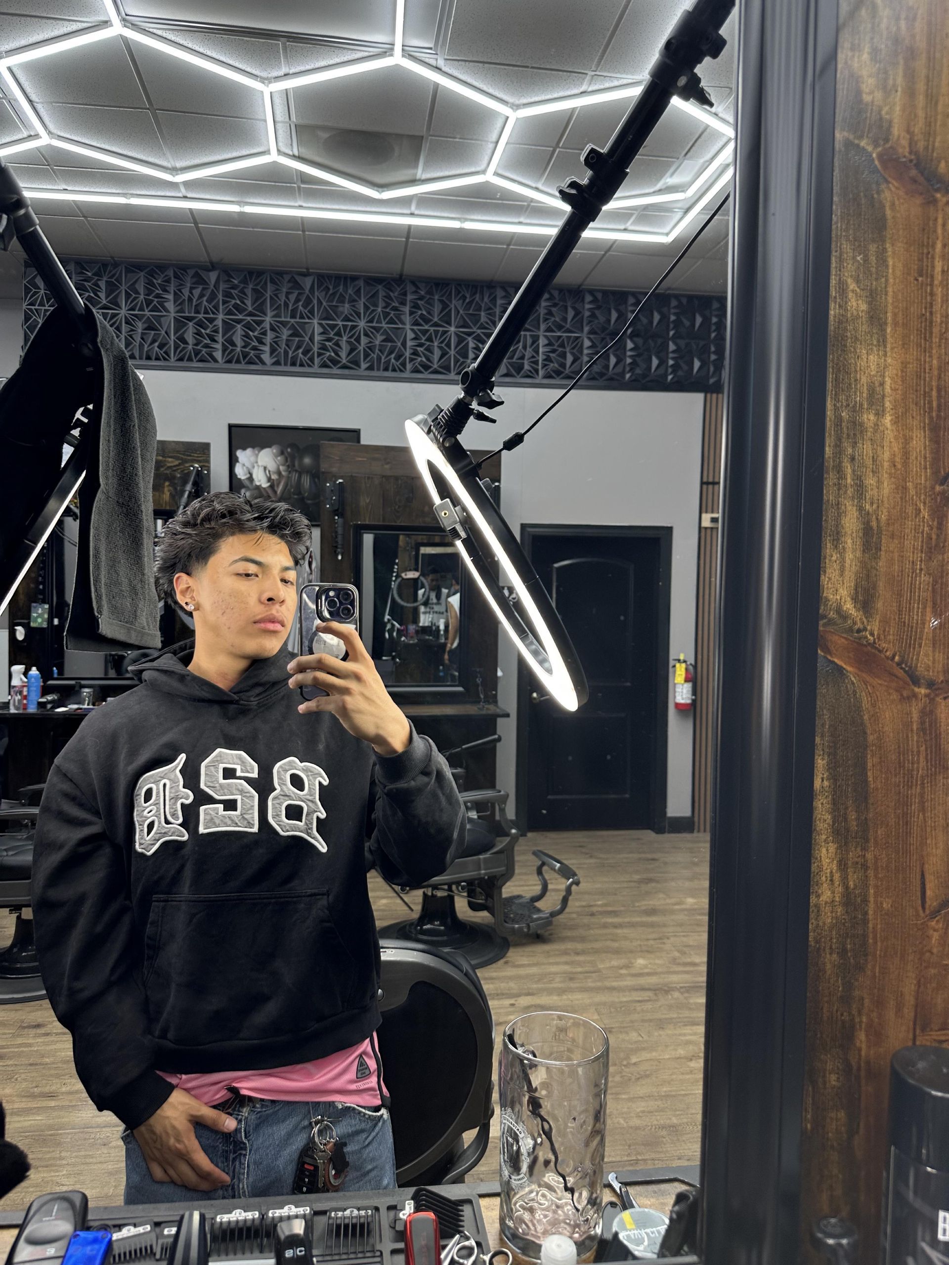 Man in a black hoodie taking a mirror selfie in a barbershop; ring light overhead.
