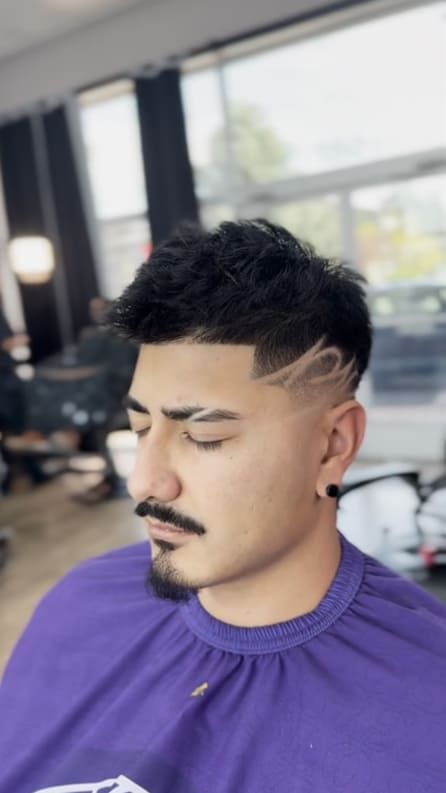 Man with a textured haircut and design, fading into a clean shave, wearing a purple barber cape.