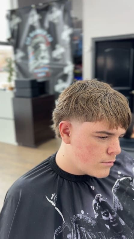 Young person with a faded mullet haircut in a barber shop, wearing a black cape.