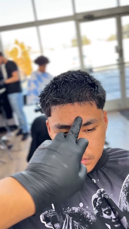 A barber with black gloves touches a man's forehead at a salon. Other people are visible in the background.