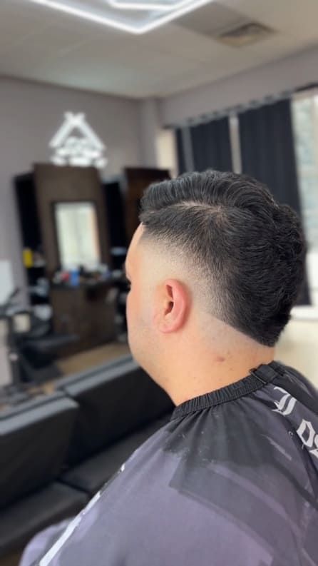 Man with dark hair, fresh fade haircut in a barber shop, wearing a black cape.