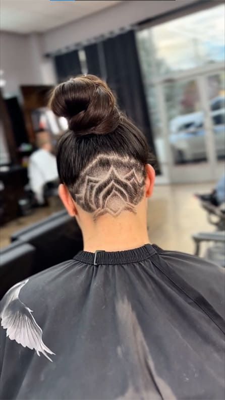 Person with intricate shaved hair design in the back, dark bun on top. Indoors, in a salon.