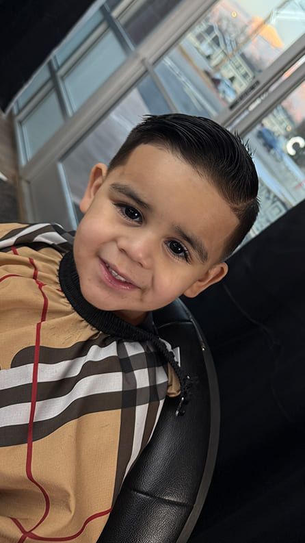 Young boy with a fresh haircut smiles at the camera in a barbershop, wearing a tan patterned cape.