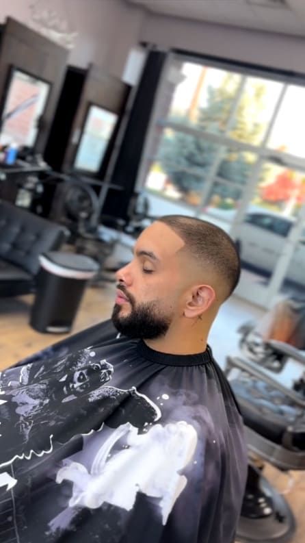Man with a faded haircut and beard in a barber shop, wearing a cape, eyes closed, in profile.