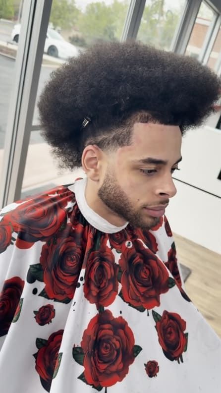 Man with an afro and beard, wearing a cape with red roses, at a barbershop.