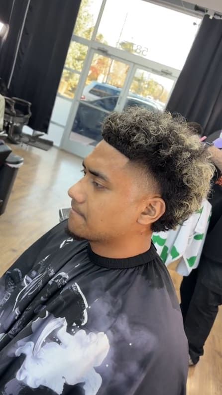 Man getting a haircut. He has a fade with curly, highlighted hair, sitting in a salon chair.