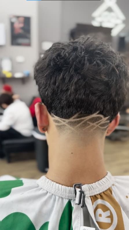 Man's back of head with dark curly hair and shaved geometric design; in a barbershop.