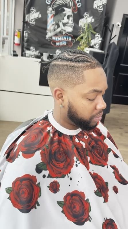 Man with braids and fade, in a barber shop, wearing a rose-print cape, getting a haircut.