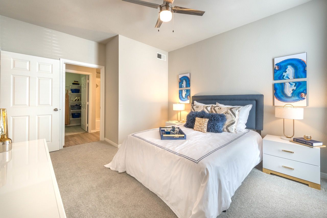 Bedroom in an apartment with a white comforter, blue upholstered headboard, two nightstands, and blue abstract art.