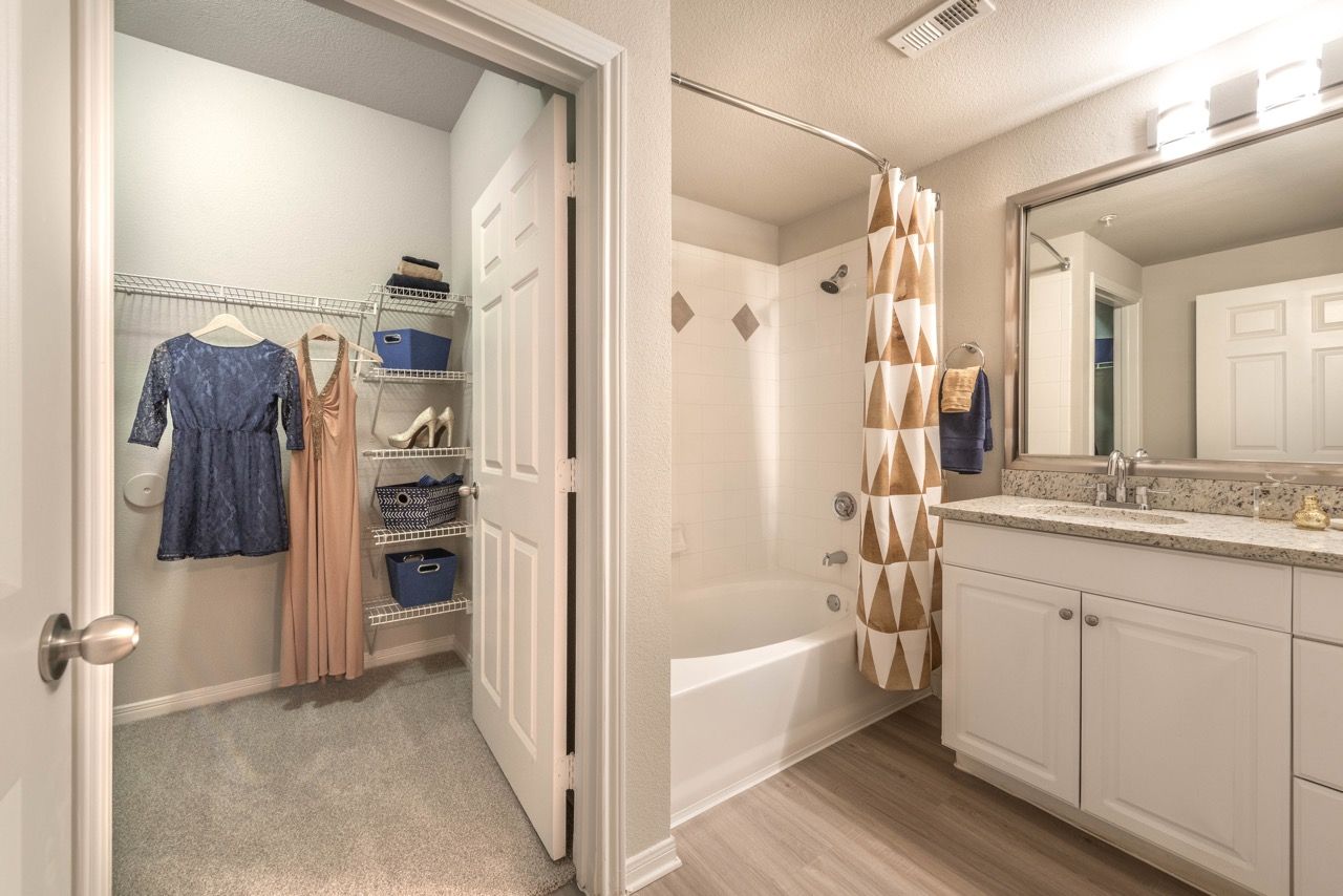 Open walk-in closet with clothes and shelves beside a bathroom tub/shower.