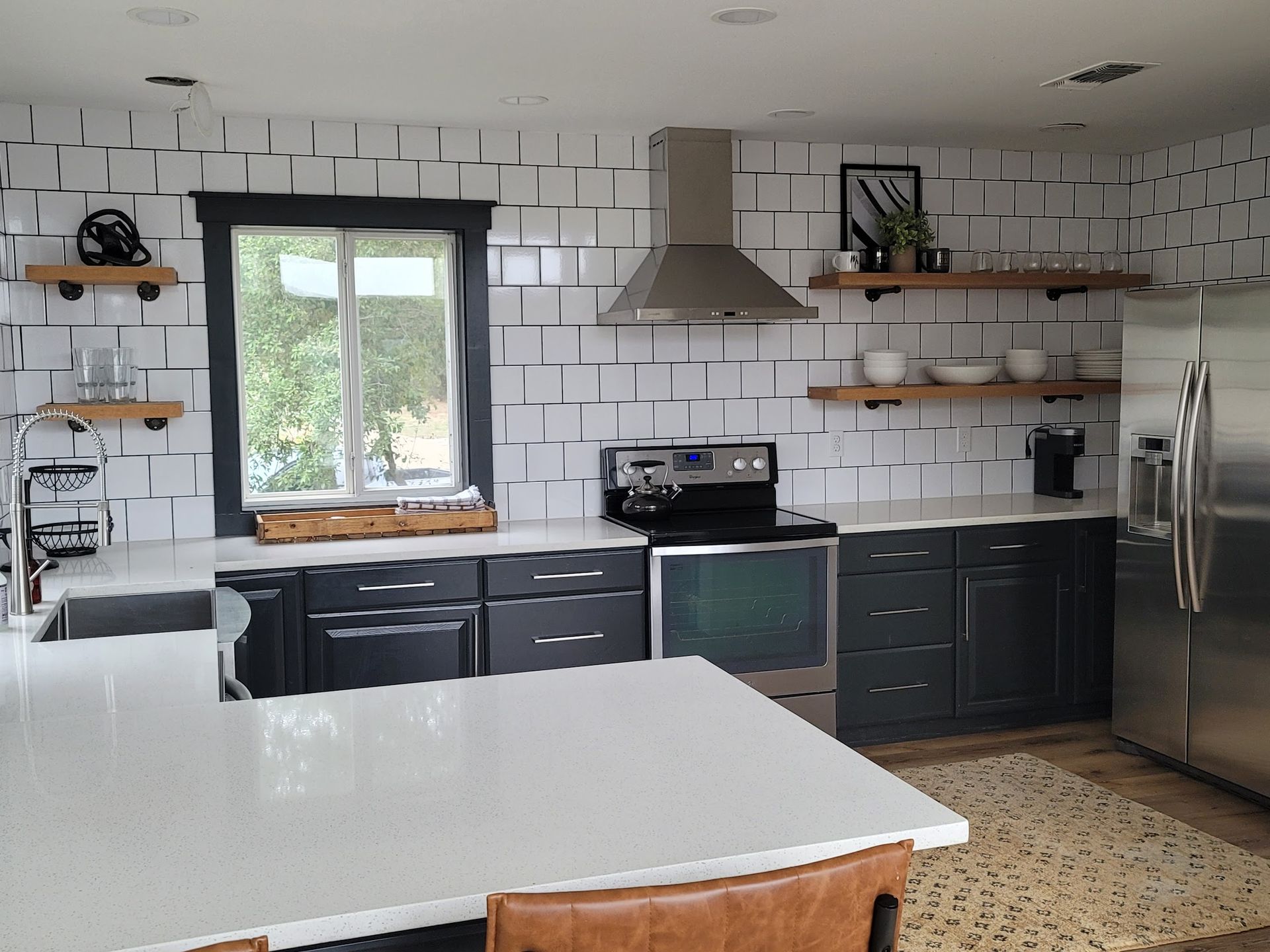 A kitchen with black cabinets , white counter tops , stainless steel appliances and a large window.