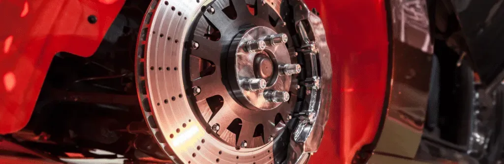 A close-up of a red car's brake system. The brake rotor is drilled and slotted with silver and black details.