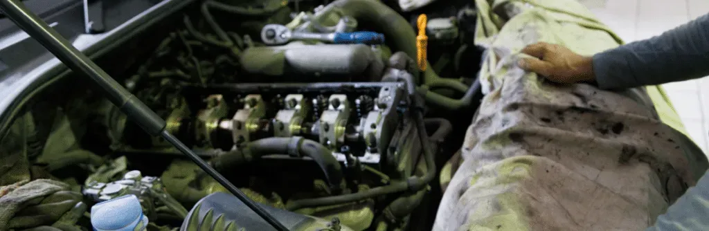 A mechanic working on a car engine. The engine bay is open, a hand is wiping the engine with a cloth.