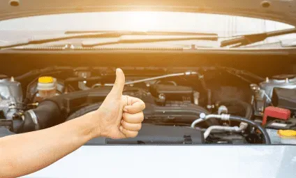 Hand giving a thumbs up in front of a car engine, suggesting approval or satisfaction.