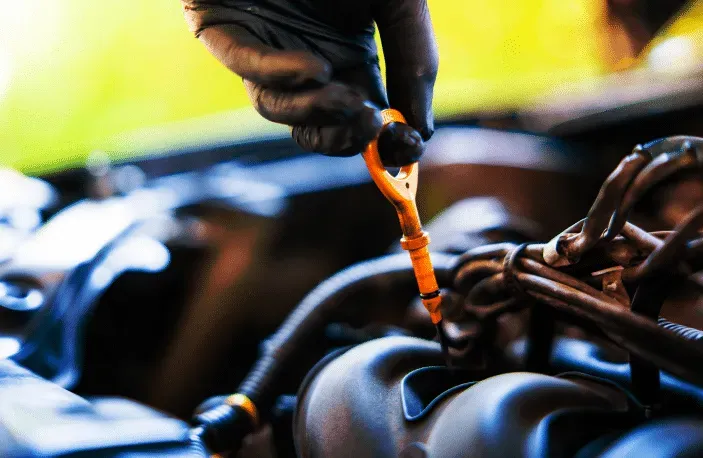 Gloved hand pulling an orange dipstick from a car engine, checking oil level in an automotive setting.