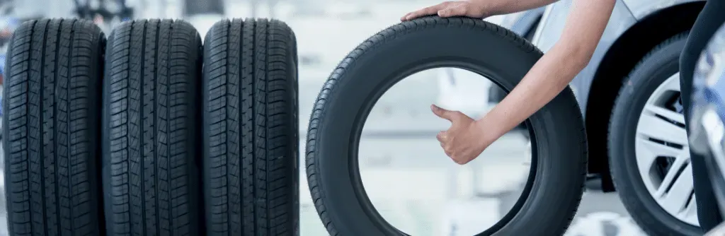 Person holding a tire in a tire shop next to other tires and a car.