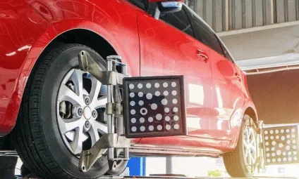 Red car undergoing wheel alignment with diagnostic equipment attached to the wheels in a repair shop.