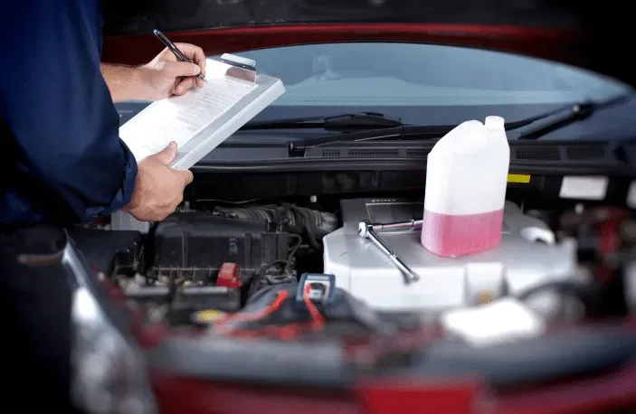 Mechanic inspecting car engine, writing on clipboard, with coolant jug nearby.