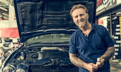 Mechanic with a beard smiles at the camera while standing next to a car with the hood open in a garage.