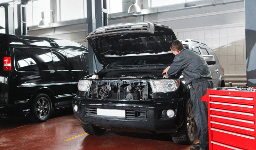 Mechanic working on a black car with its hood open inside a garage, next to a red toolbox.