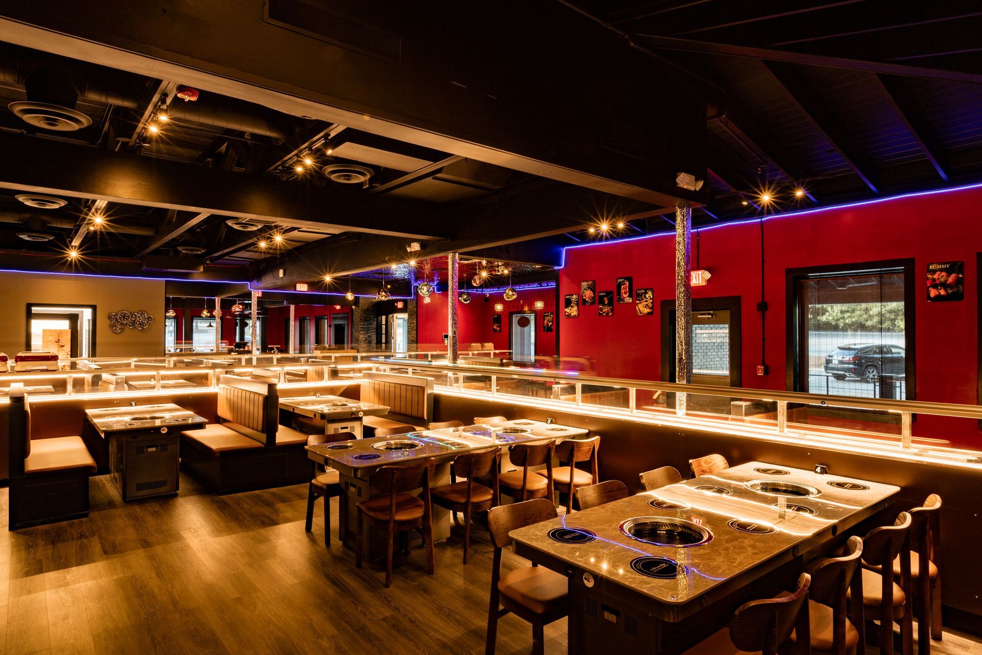 Interior of a Korean barbecue restaurant with red walls, dark tables, and built-in cooking surfaces. Tables are arranged with booths and chairs.