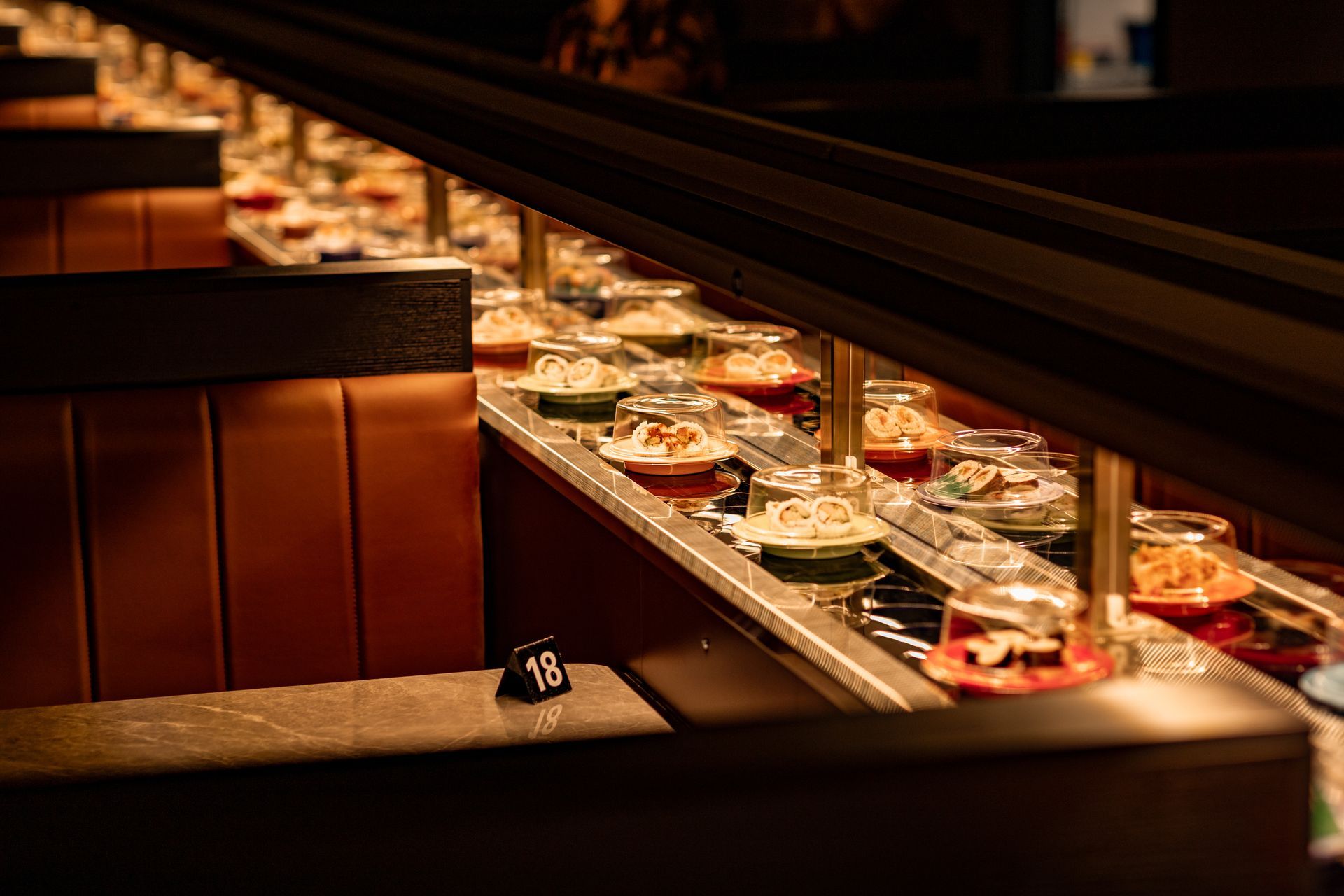 A conveyor belt sushi restaurant interior with small dishes of food traveling past booths. Number 