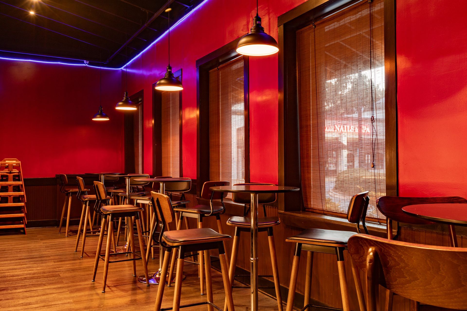 A dimly lit interior of a bar/restaurant with red walls, tables, stools, and hanging lights near windows.