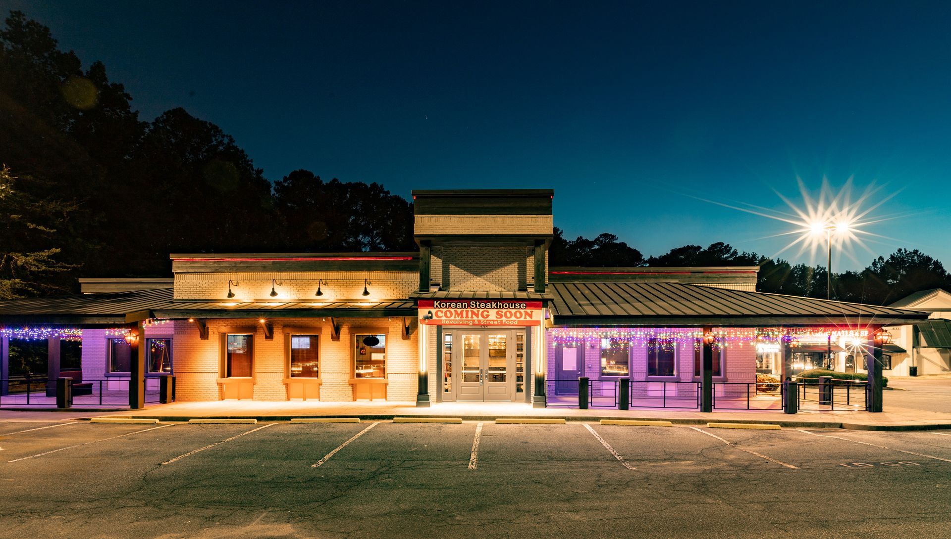 A brightly lit restaurant exterior at night, with colorful accent lighting, parking lot in front, and a dark night sky.