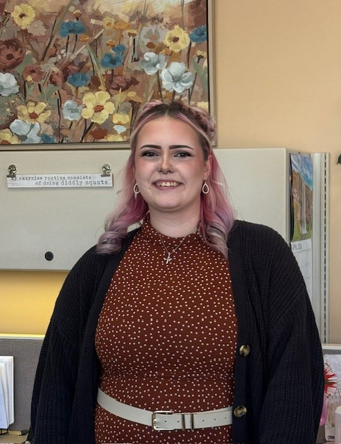 Woman standing in an office, smiling, with pink hair, burgundy dress, and dark cardigan.