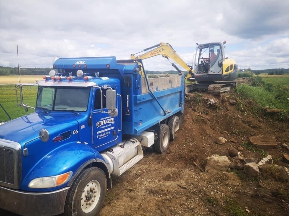 Un camion-benne bleu roule sur un chemin de terre à côté d’une excavatrice.
