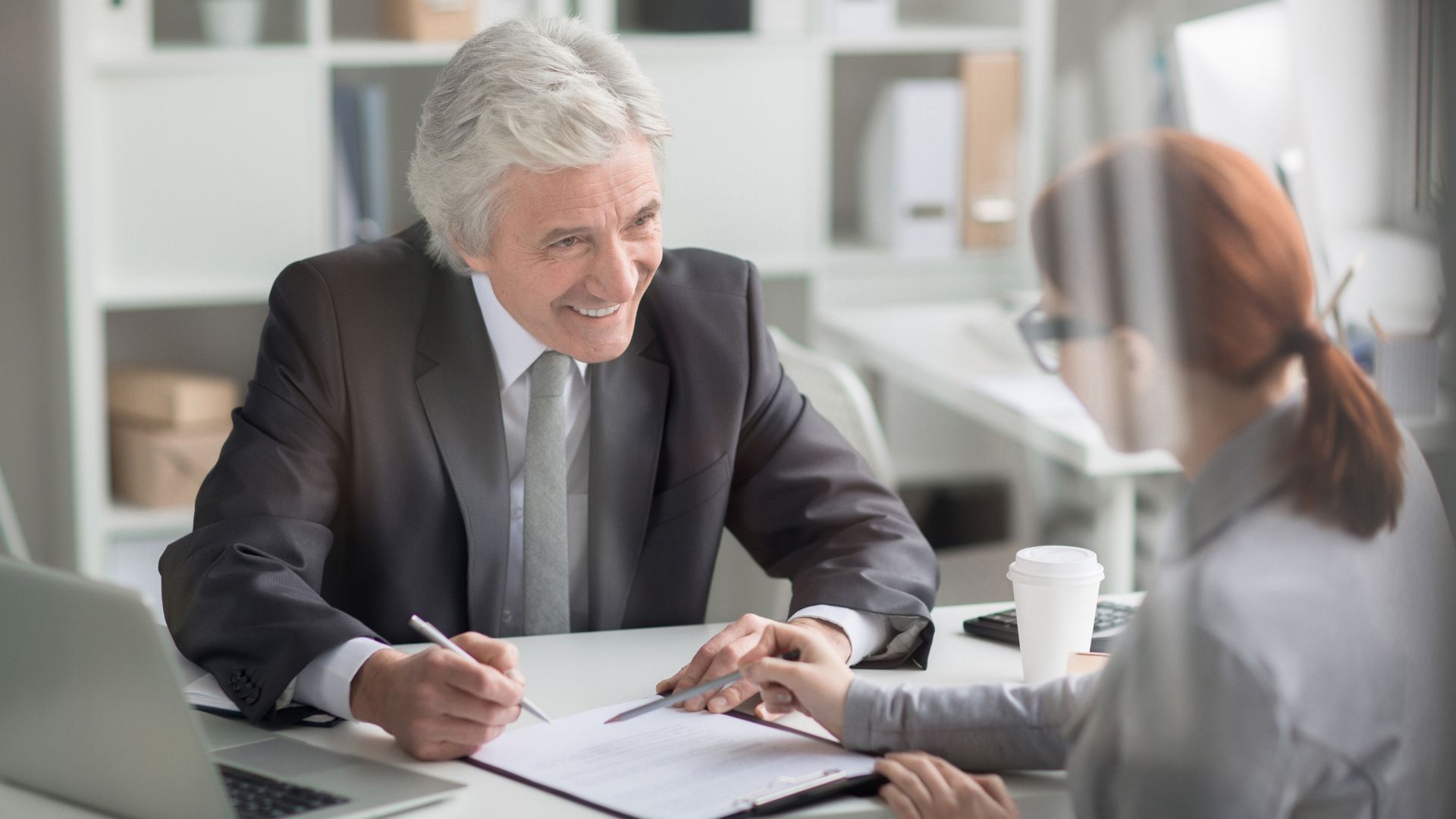 Man in suit smiles, signing document with a woman at an office desk.