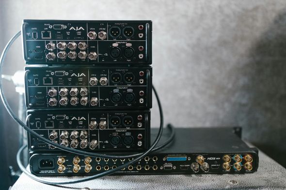 Four black AJA video converters stacked on top of each other, next to a black rackmount device, on a grey surface.