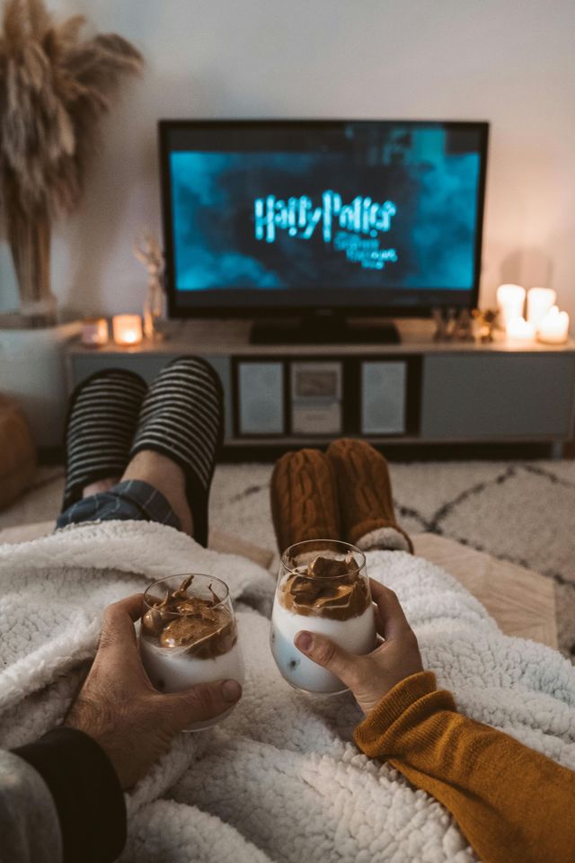 Couple relaxing, watching Harry Potter with whipped coffee and cozy blankets, soft lighting.