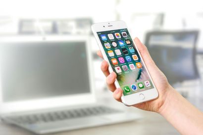 Person holding white smartphone with app icons, near a laptop on a desk.