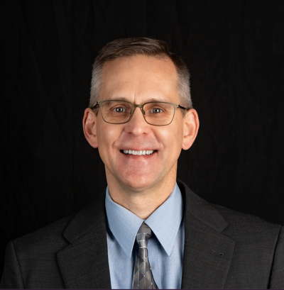 A man in a plaid shirt is smiling in front of a wooden wall.