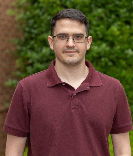 A man wearing glasses and a maroon polo shirt is standing in front of a tree.