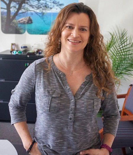 A woman in a grey shirt is standing in a room with her hands in her pockets.